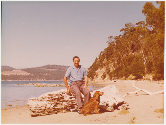 Original, photo with faded colors. Photo of a scenic sandy beach showing an adult male sitting on a dry log, with his large dog sitting nearby.
