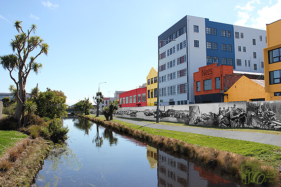 Image of buildings next to a disused strip of land which is situated between a long straight canal and a long concrete wall is now transformed into a picturesque walkway with the addition of plants and the concrete wall now decorated with historical photographs/murals.