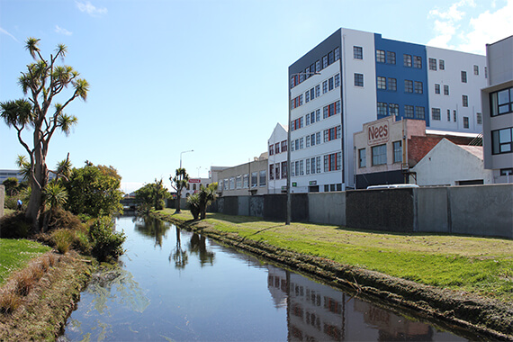 Image of buildings next to a disused strip of land which is situated between a long straight canal and a long concrete wall.